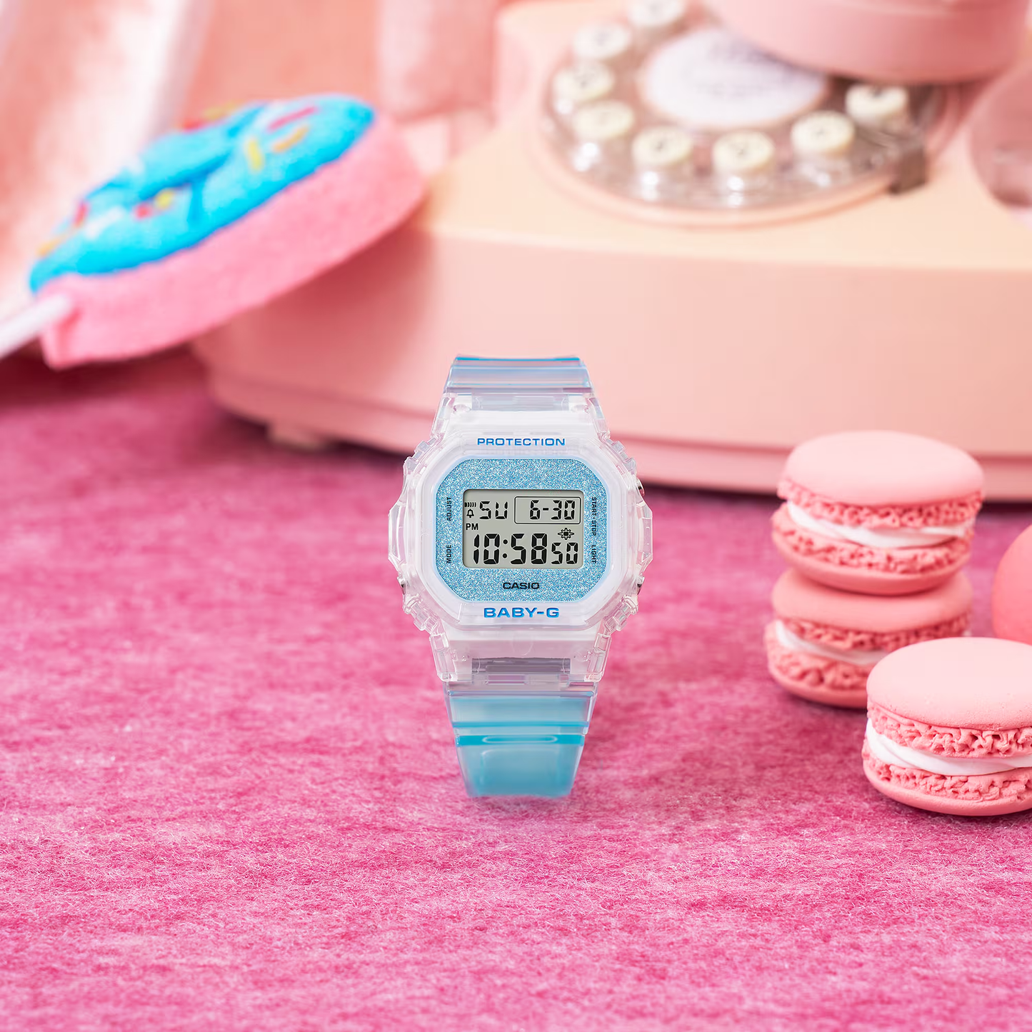 Baby-G digital watch with blue strap on a pink surface with macarons and a phone in the background