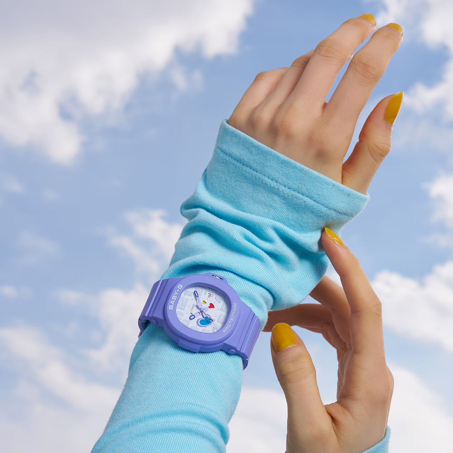 Hand wearing a blue wristband with a purple watch against a blue sky.