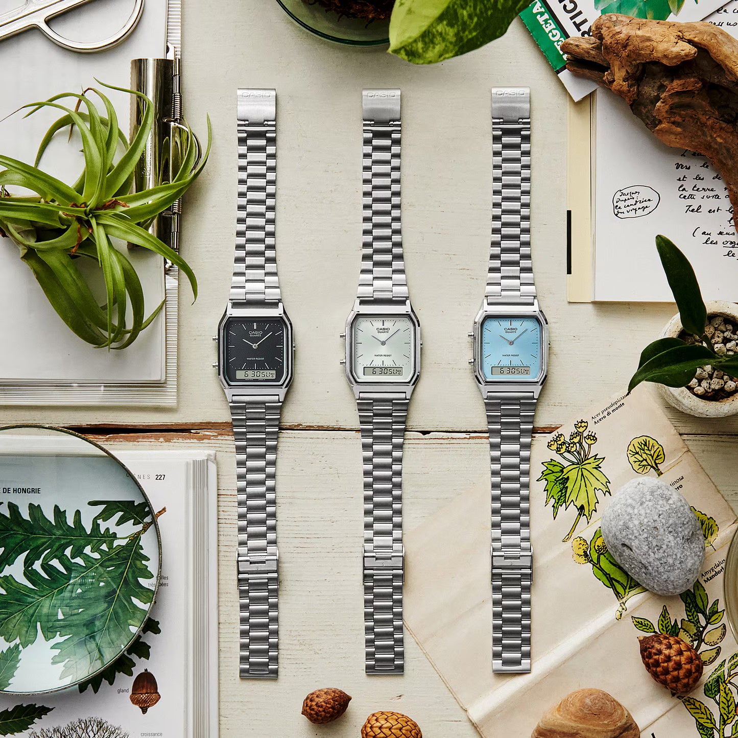Three silver watches on a decorative surface with plants and nature-themed items.