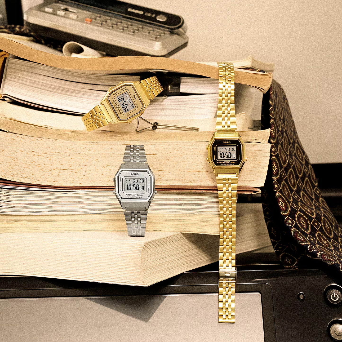 Three digital watches on a stack of books with a vintage computer in the background.
