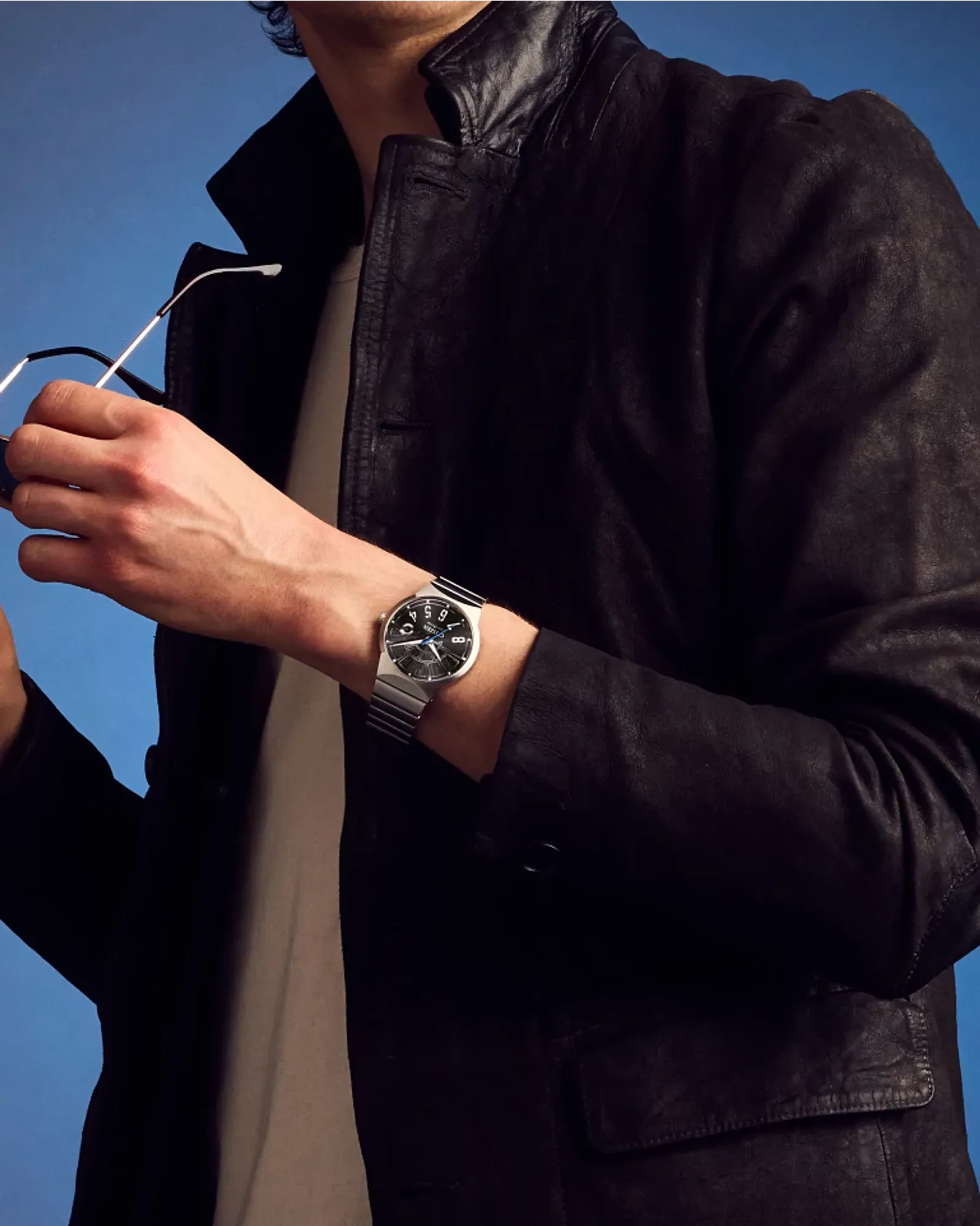 Person wearing a watch with a dark jacket against a blue background
