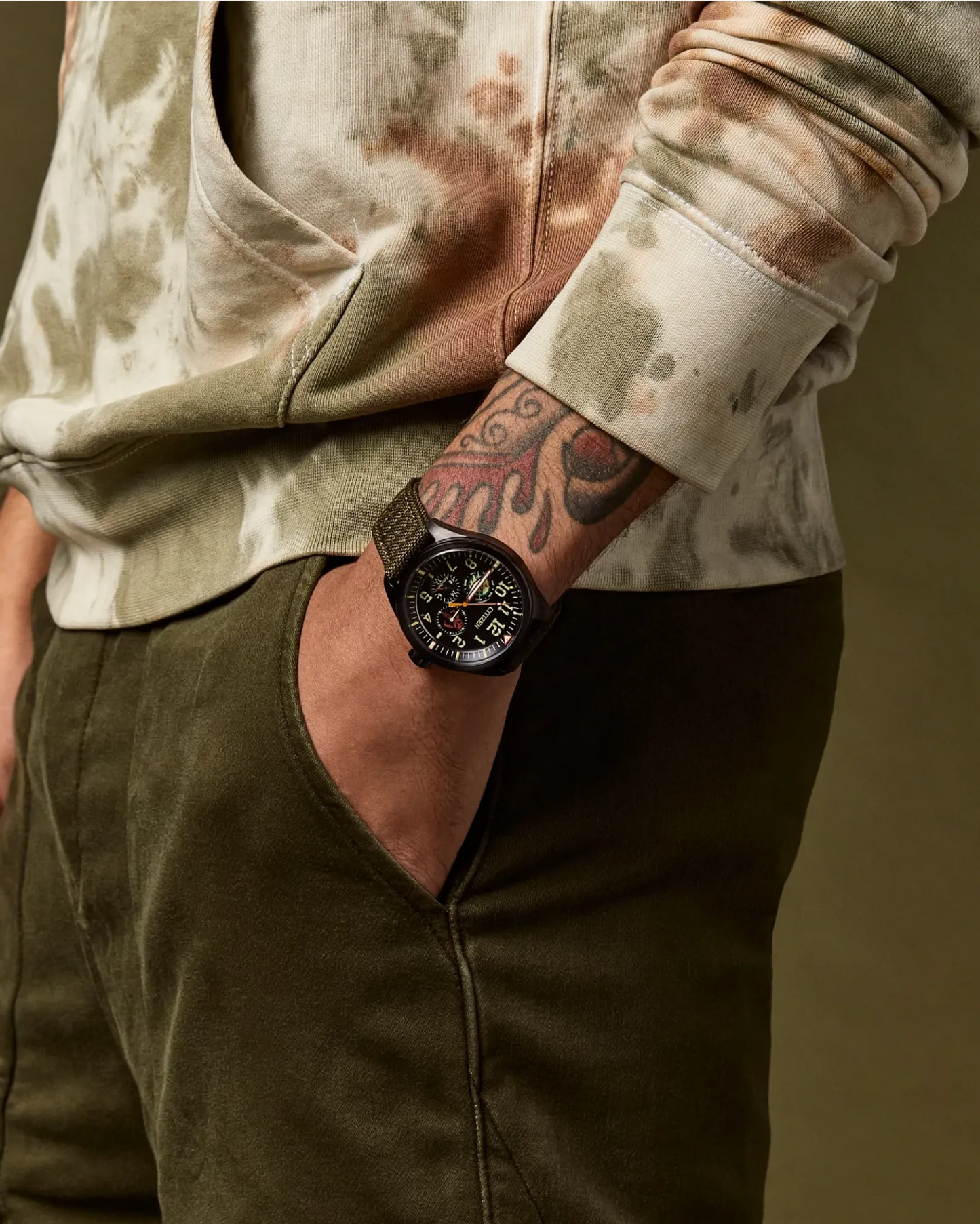 Person wearing a camouflage jacket and green pants with a tattooed arm, holding a watch.