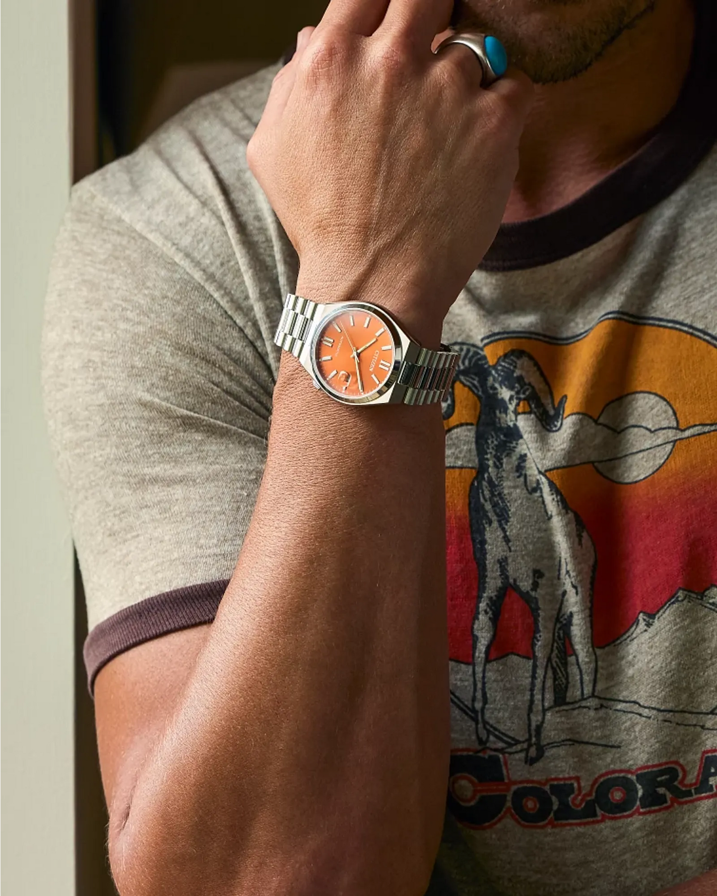 Person wearing a watch with an orange face, gray case, and silver band, on a gray t-shirt with a graphic design.