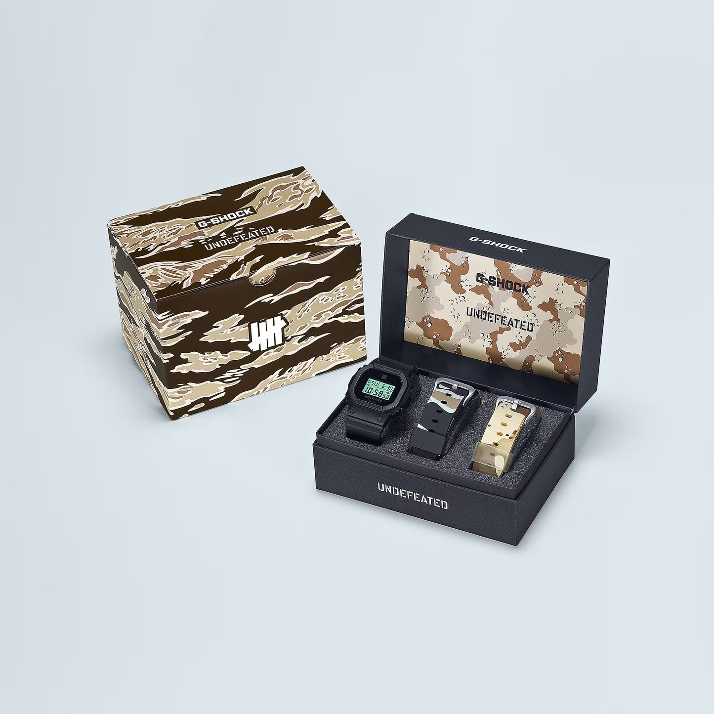 Casio G-SHOCK DWE5600UD-1 UNDEFEATED collaboration model with matte black Carbon Core Guard case, semi-translucent resin band featuring UNDEFEATED logo, green LCD display, engraved caseback, and special packaging. Includes two interchangeable camouflage straps (tiger camo, storm camo). Features shock resistance, mineral glass, 200m water resistance, LED Super Illuminator backlight, stopwatch, countdown timer, alarms, flash alert, auto-calendar, and ±15 sec/month accuracy. Case size 48.9 × 43.8 × 13.7 mm, we