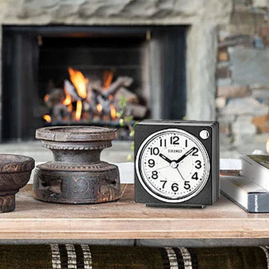 SEIKO QHE196KLH black compact bedside alarm clock displayed near a fireplace, featuring a white analog dial and easy-to-read numerals.