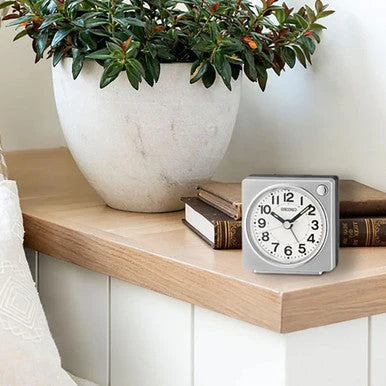 SEIKO QHE196SLH compact square alarm clock displayed on a bedside shelf with easy-to-read analog dial, silver case, and LumiBrite-tipped hands.