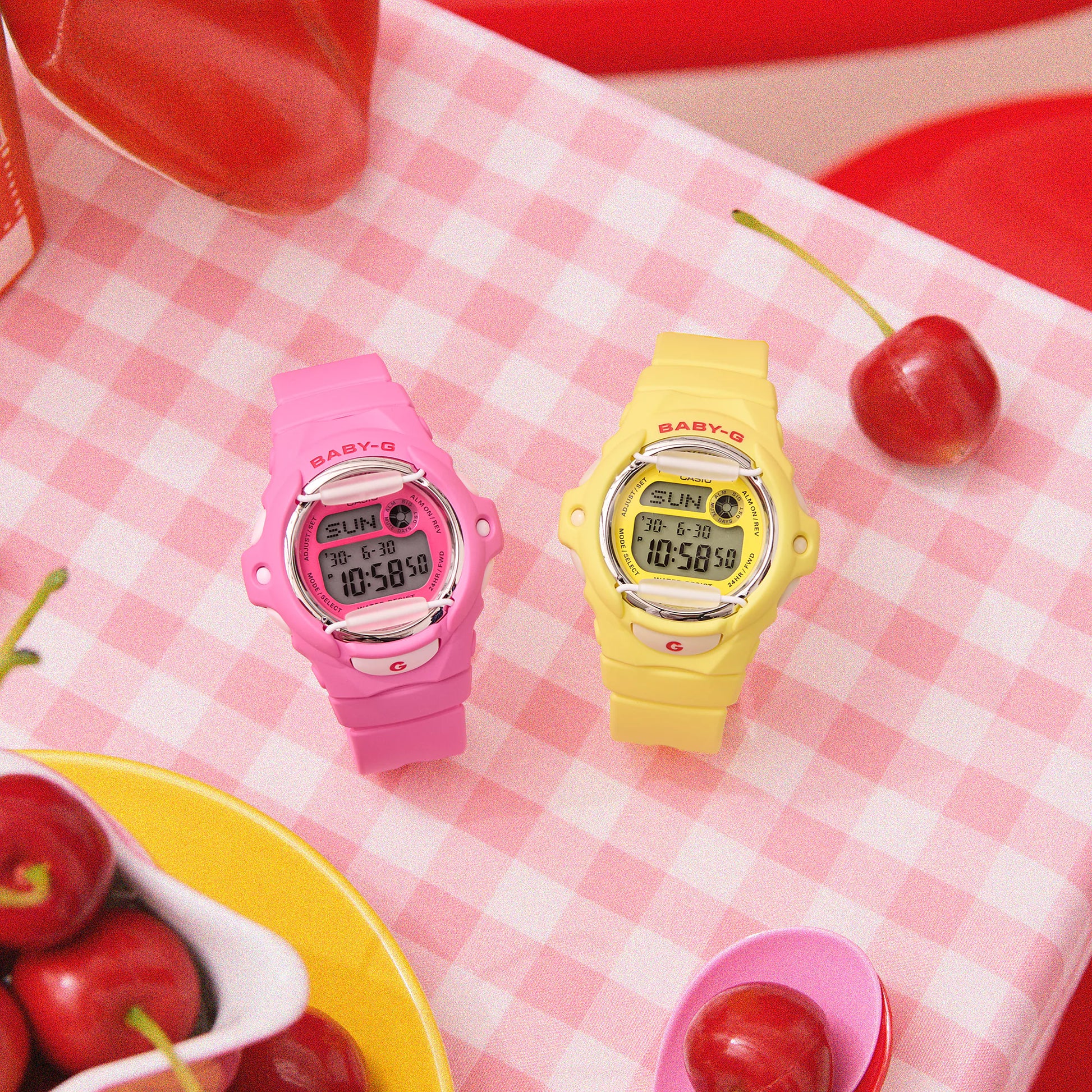 Two colorful digital watches on a pink checkered tablecloth with cherries.