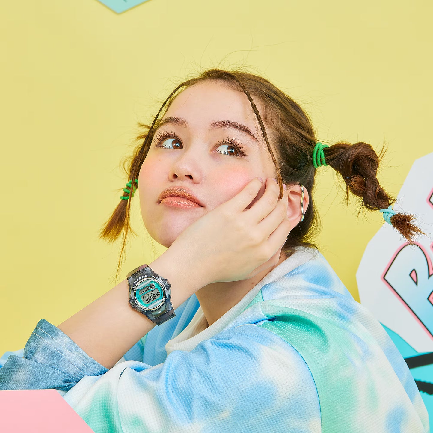 Young girl with pigtails wearing a watch, looking up with a thoughtful expression on a yellow background.