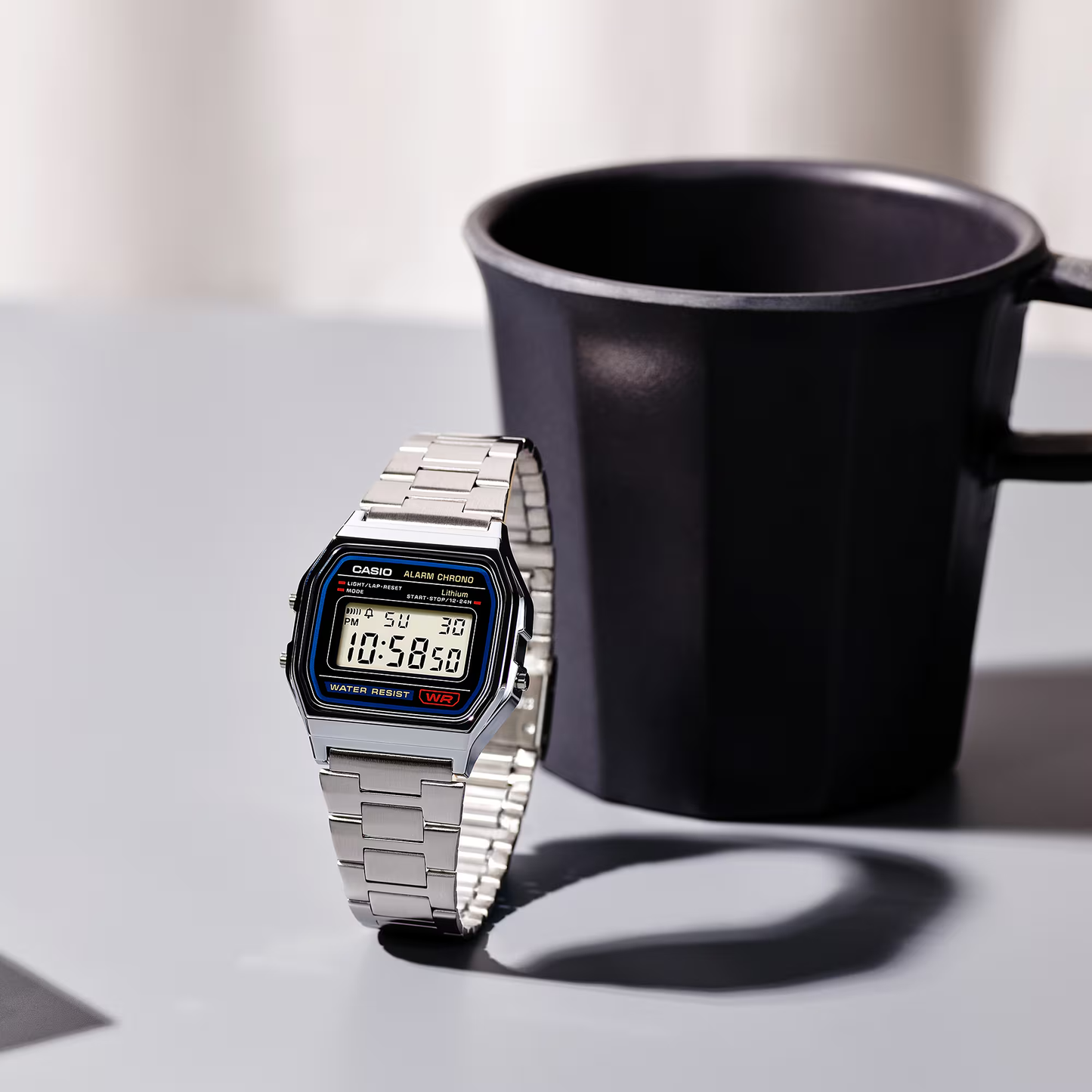 Silver Casio digital watch next to a black mug on a light surface