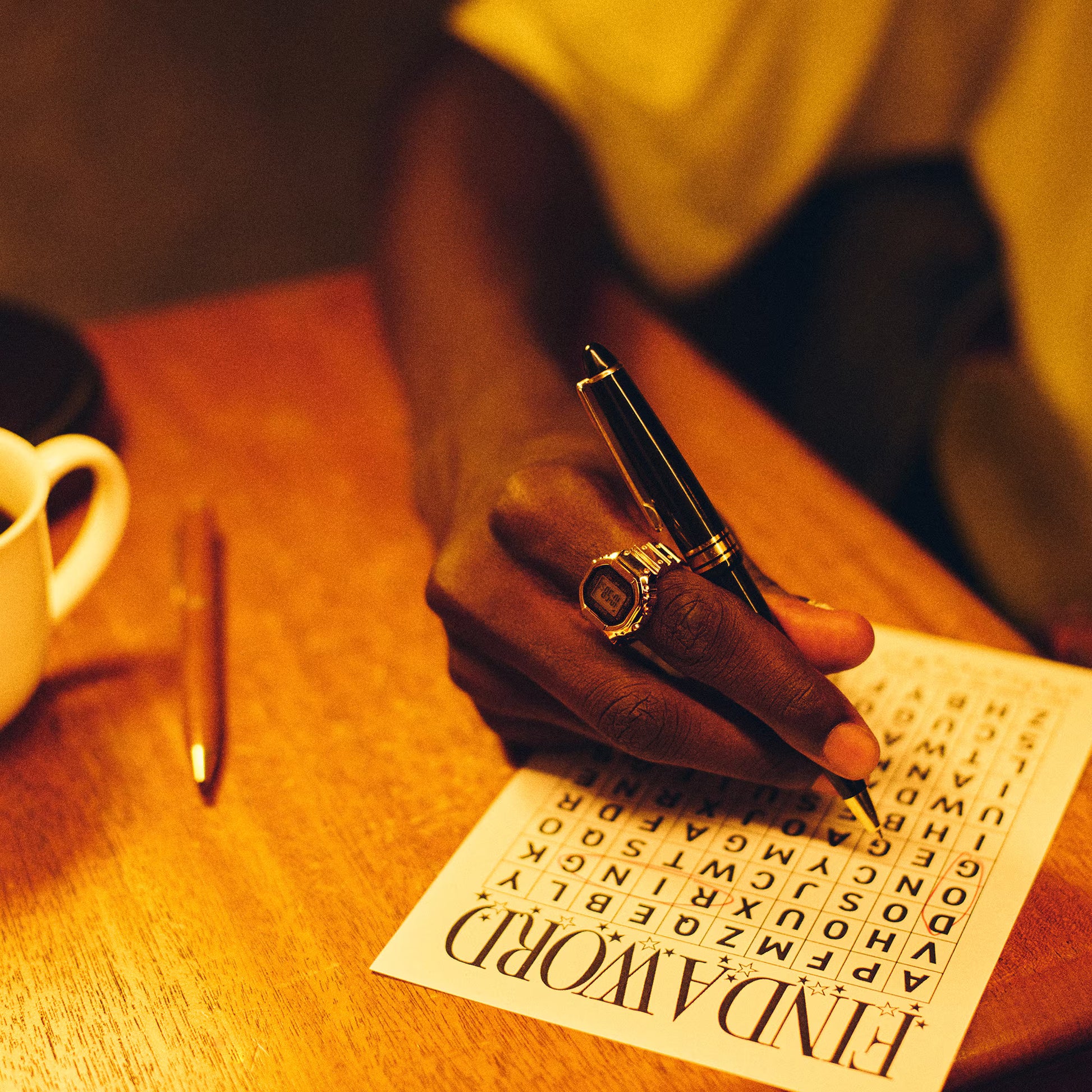Person holding a pen over a crossword puzzle on a wooden table with a cup and another pen in the background.