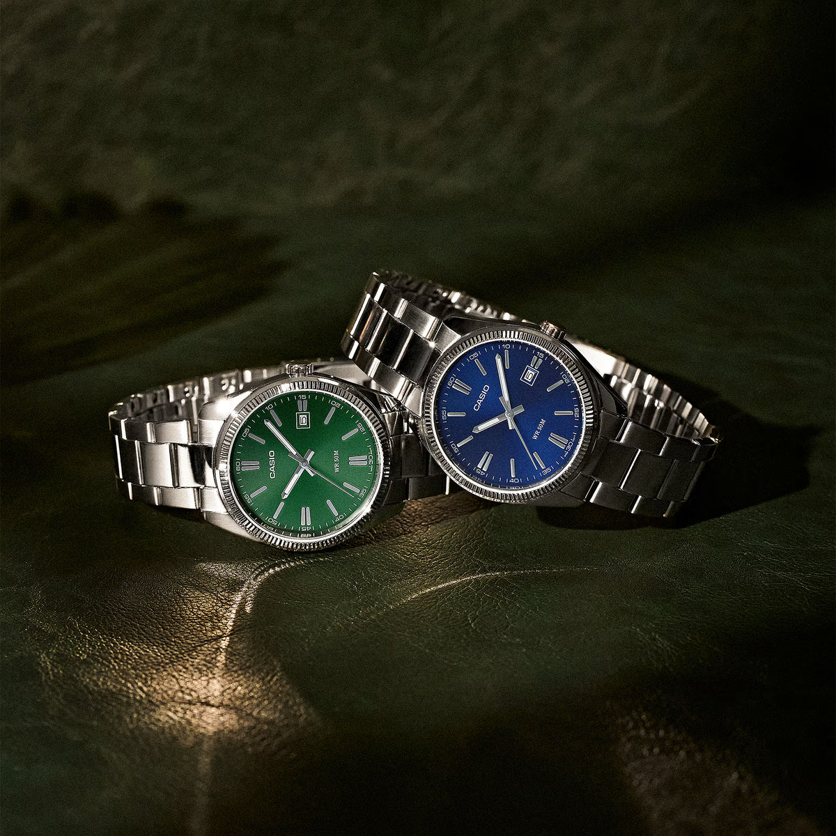 CASIO MTP1302D-2AVT blue dial and CASIO green dial stainless steel analog watches displayed together on a dark leather surface.
