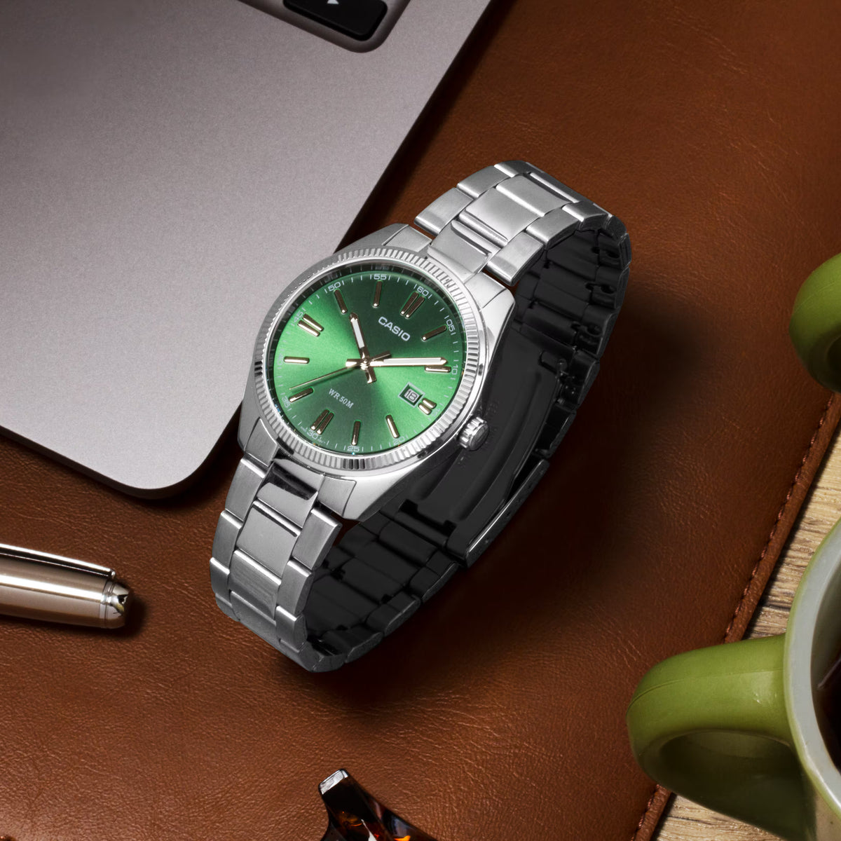 CASIO MTP1302D-3AVT green dial stainless steel watch styled on a desk with a laptop, pen, leather mat, and green coffee cup.