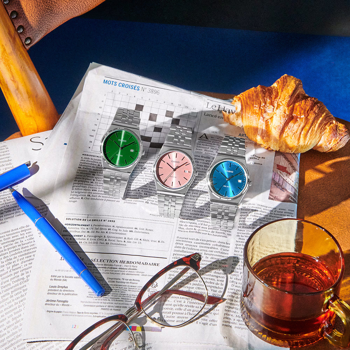 CASIO MTPB145DC-3AV green dial stainless steel watch shown with pink and blue dial variants on a newspaper beside a croissant, tea, glasses, and pen