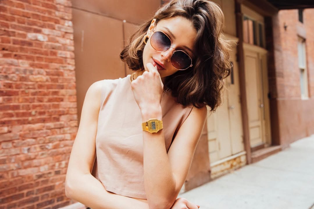 Woman wearing sunglasses and a gold watch, sitting against a brick wall.