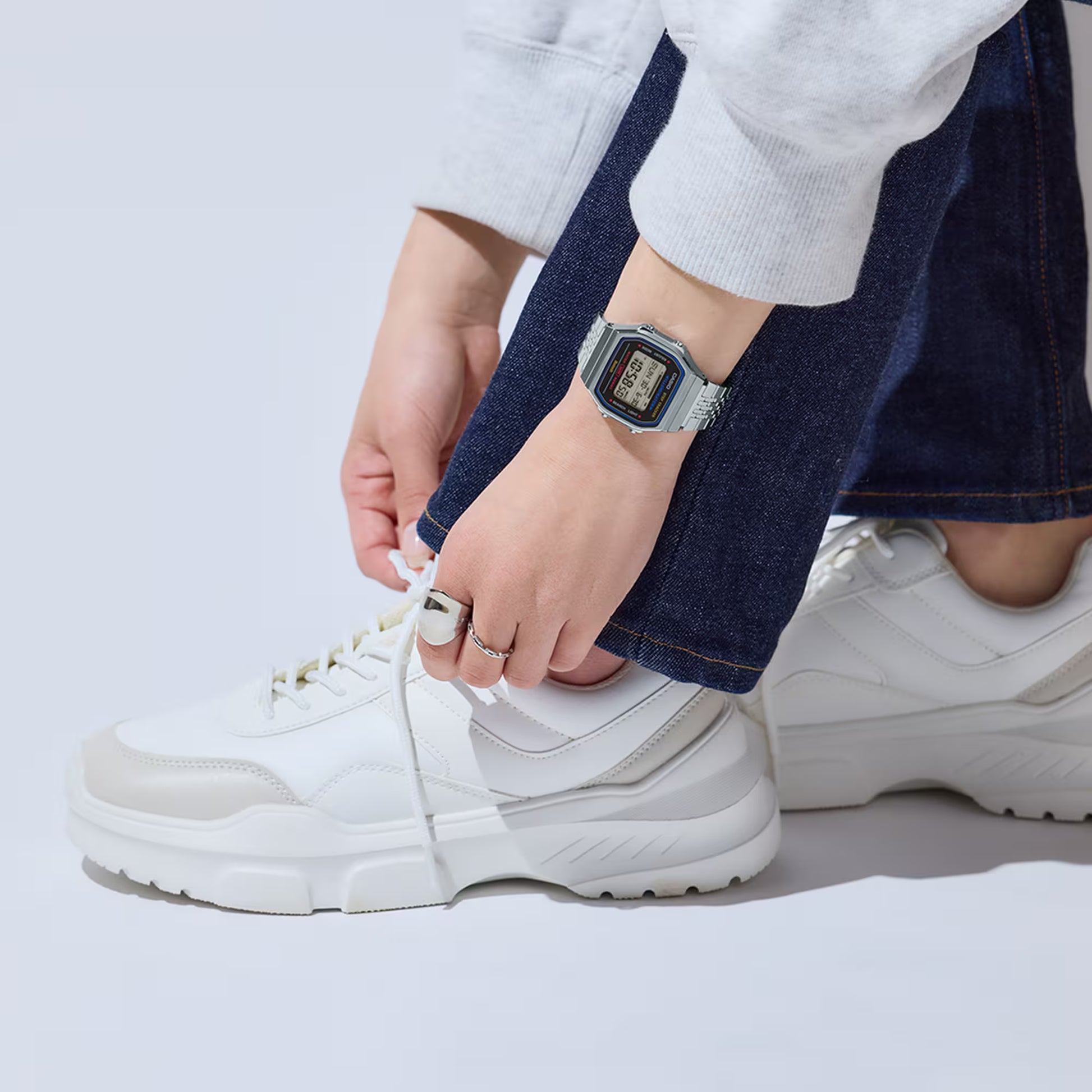 Person wearing white sneakers and a silver watch, tying shoelaces on a light gray background