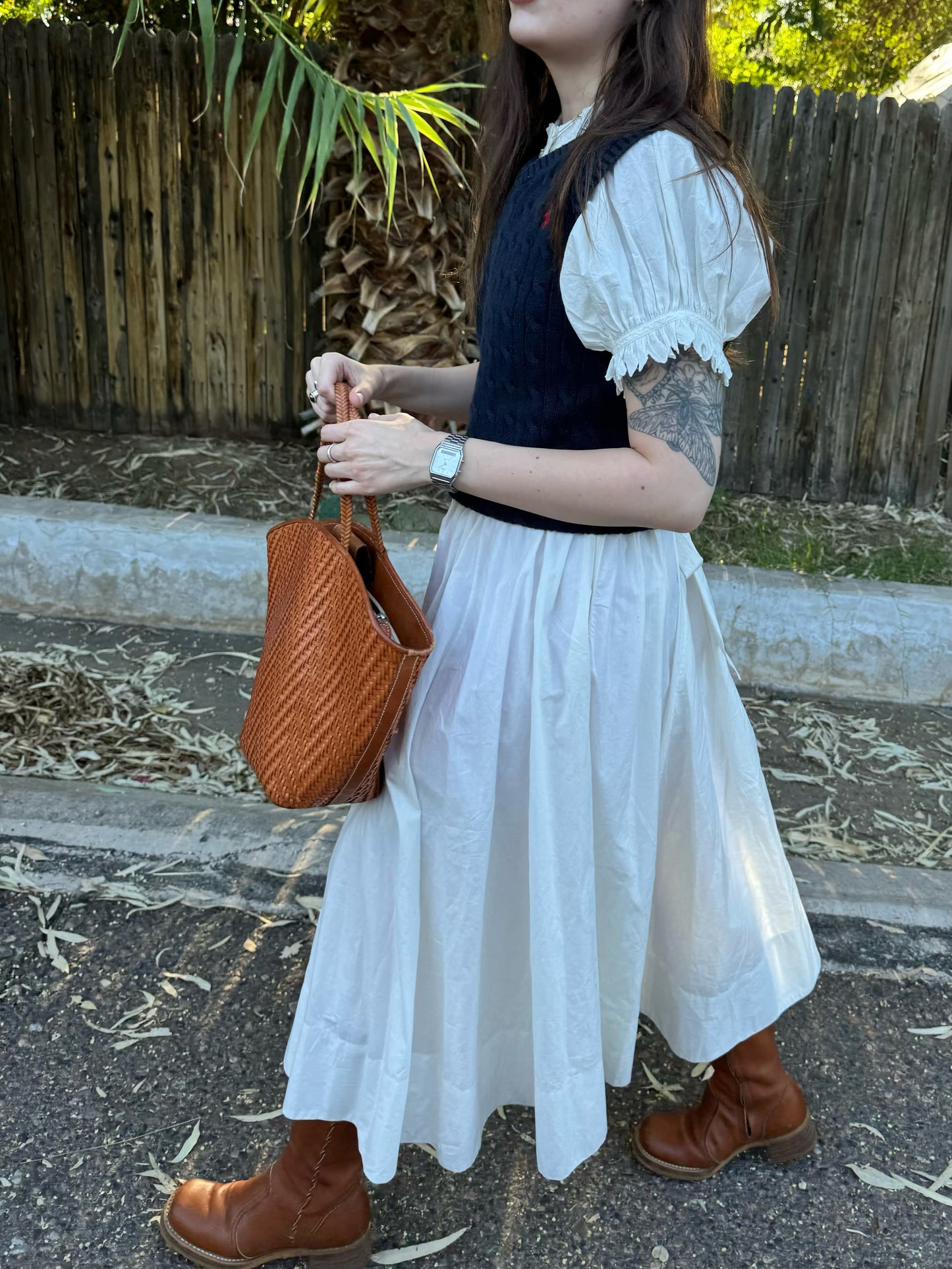 Person wearing a white dress with puffed sleeves, holding a brown woven handbag, standing on a stone path.