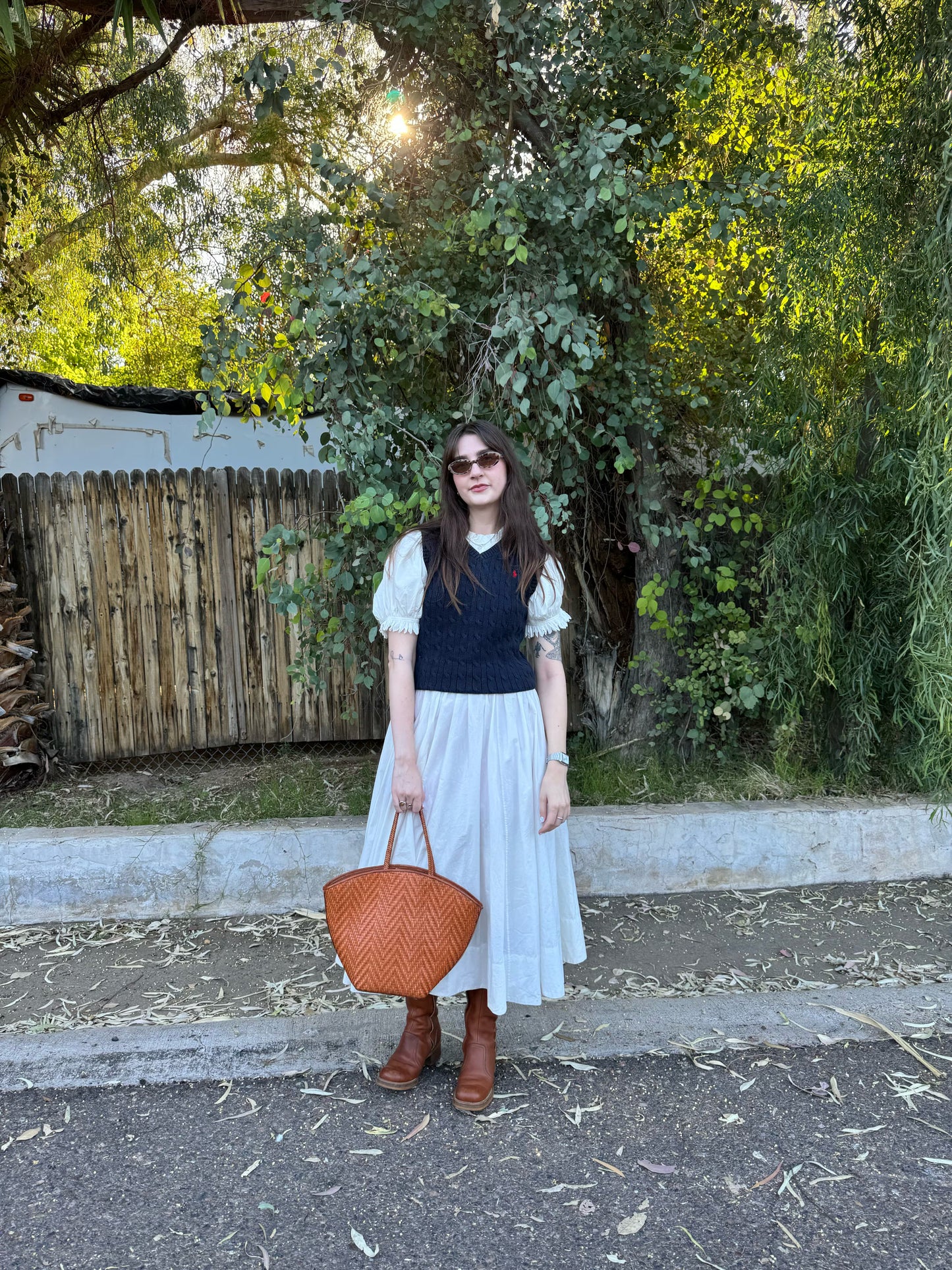 Woman standing outdoors holding a brown bag, wearing a white dress and dark top.