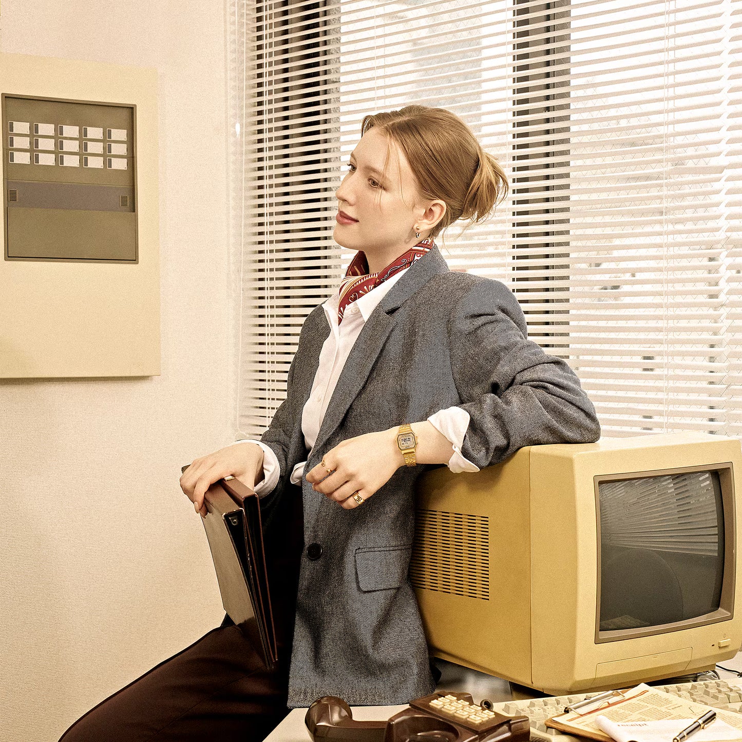 Woman in a gray suit leaning against an old computer monitor in an office setting.