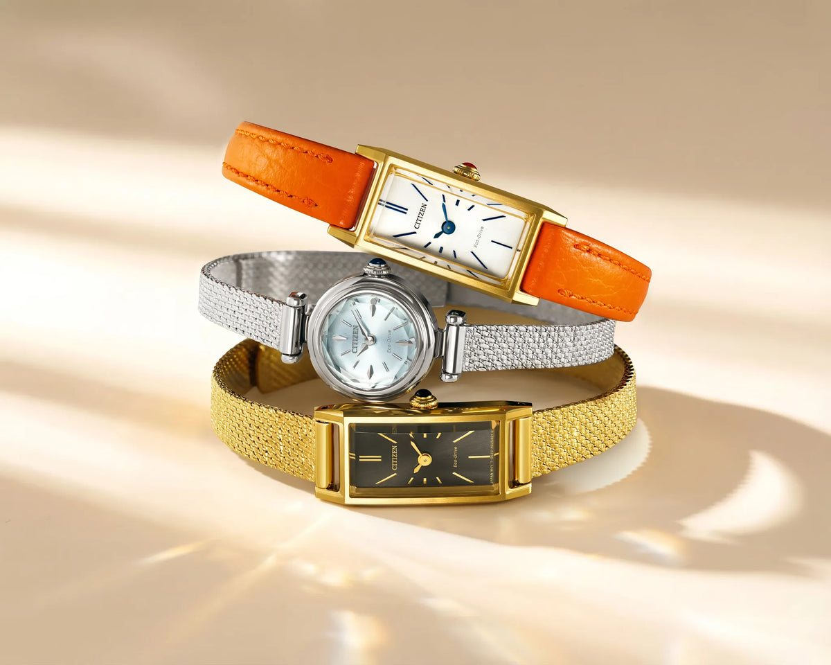 Three women's analog wristwatches stacked together on a warm beige background with soft sunlight and shadows. The top watch has an orange leather strap and gold case.