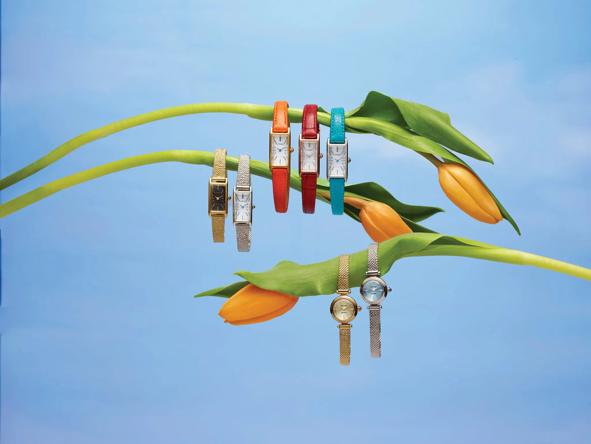 Four colorful watch straps displayed on green tulip stems against a light blue sky background. The watches feature silver-toned rectangular cases and are shown in red, turquoise, gold, and silver-tone versions.