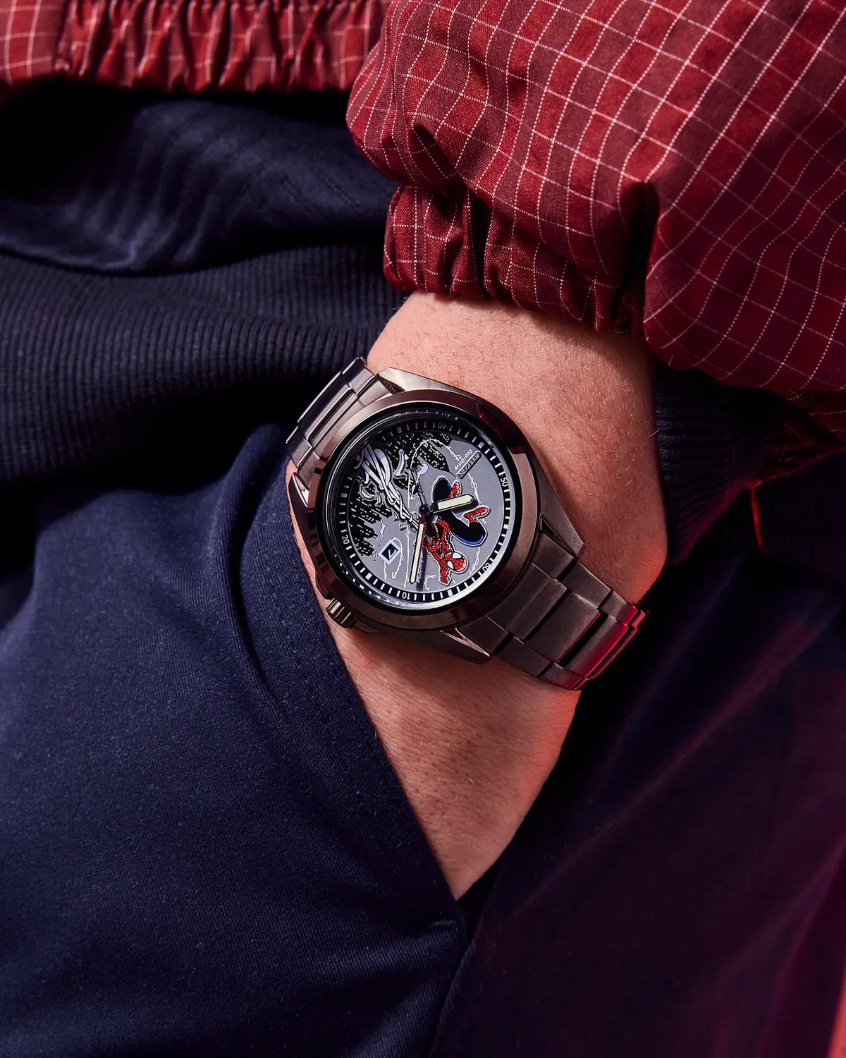 A close-up of a men's wristwatch with a dark metallic link bracelet and a detailed skeleton-style dial, worn on a person's wrist. The person is wearing a red and white plaid shirt and dark navy pants.