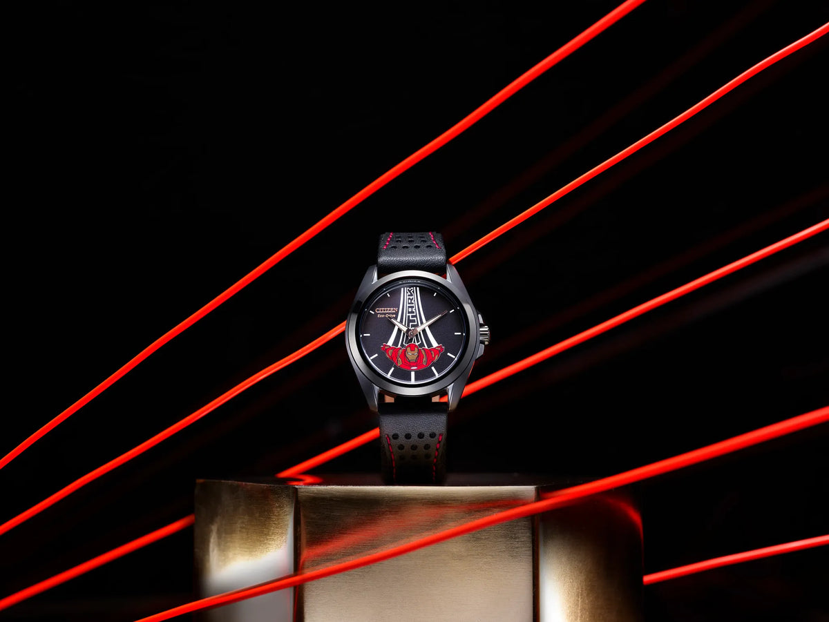 Citizen Marvel Stark AW2095-41W displayed on a metallic pedestal with red light trails, featuring Tony Stark dial artwork, black ion-plated case, and red-stitched leather strap.