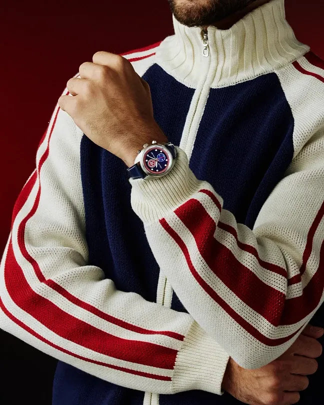 A close-up lifestyle shot of a man wearing a Gucci track jacket with navy blue, cream, and red stripes, wearing a Gucci watch on his wrist against a dark red background.
