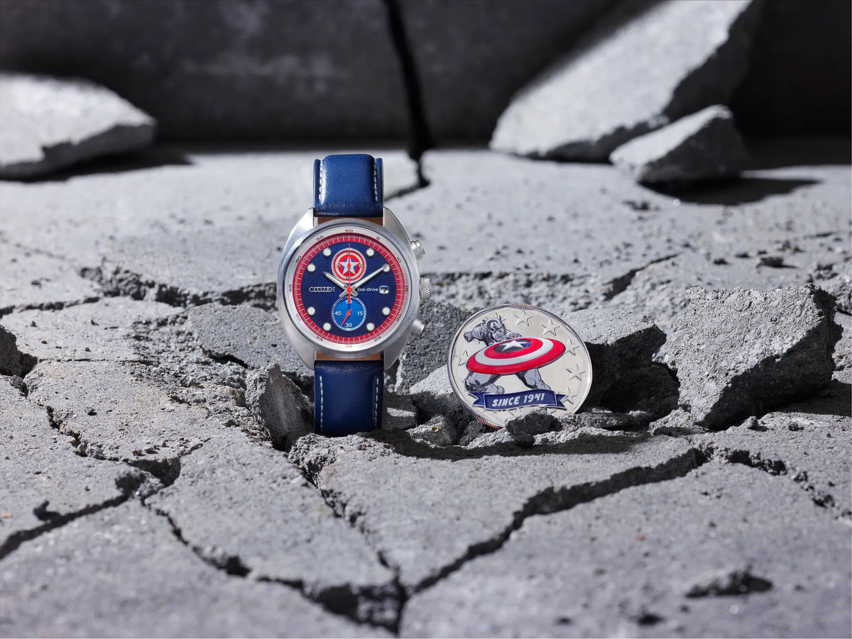 A Captain America themed watch with a blue leather strap and a red dial, displayed next to a matching Captain America medallion on a cracked gray concrete surface.