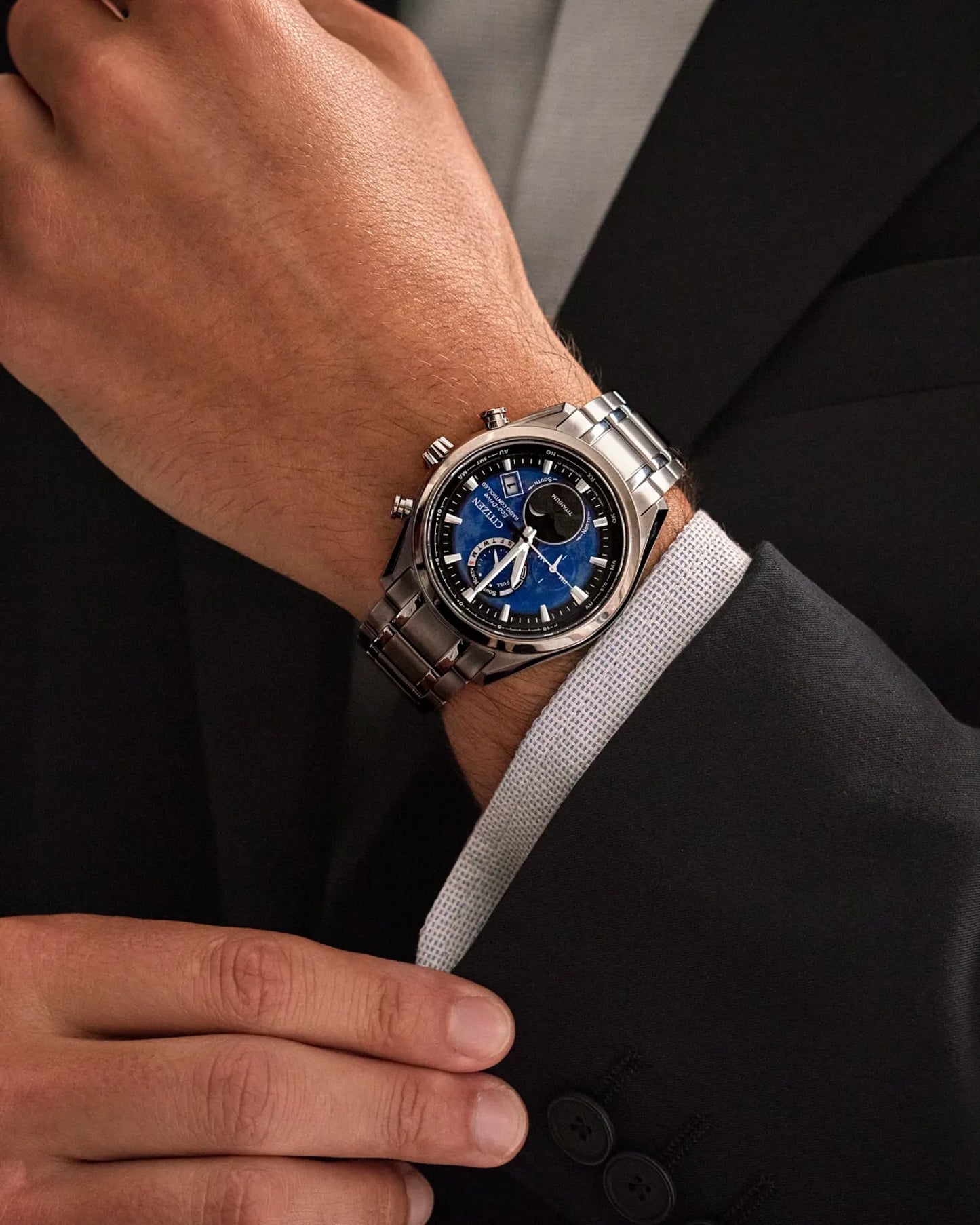 Close-up of a wrist wearing a silver watch with a blue face, against a dark suit background.