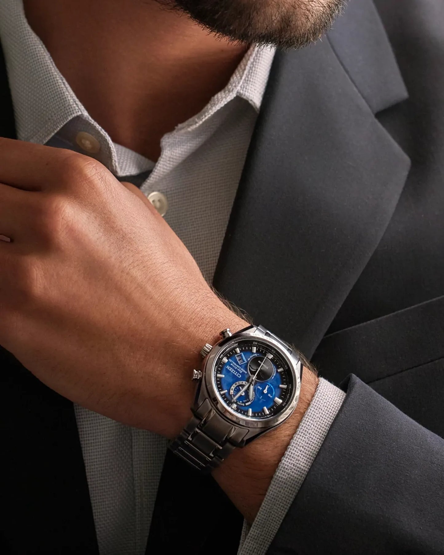 Man wearing a silver watch with a blue face on his wrist, dressed in a gray suit.