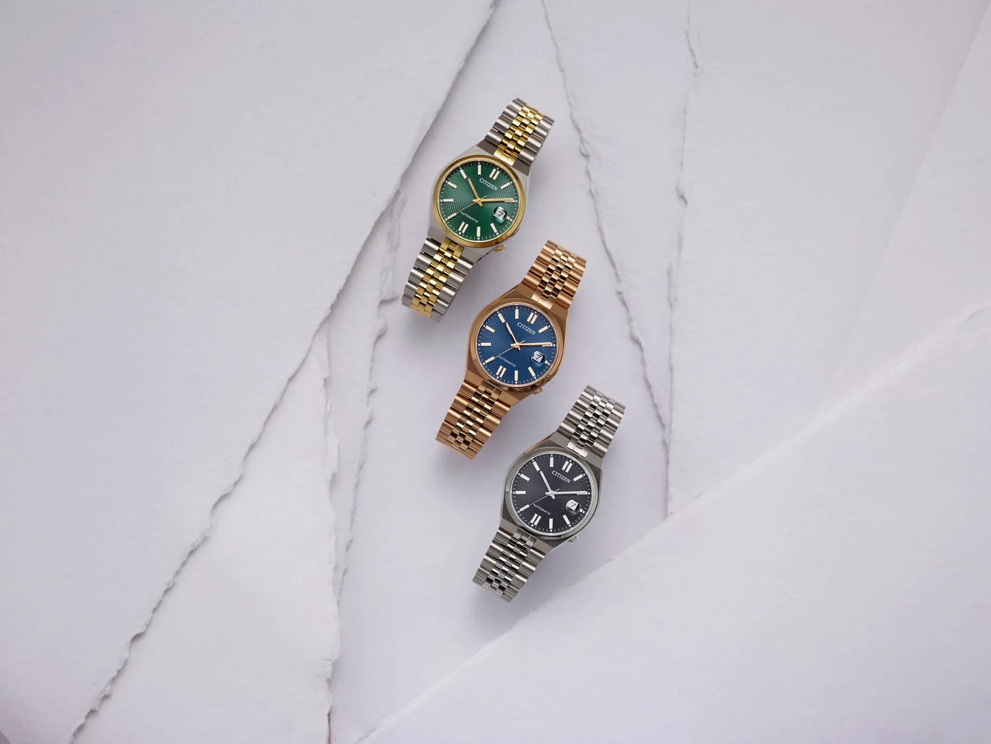 Three women's watch bracelets arranged diagonally on a white marble surface. The top watch has a gold and silver link bracelet with a green dial.