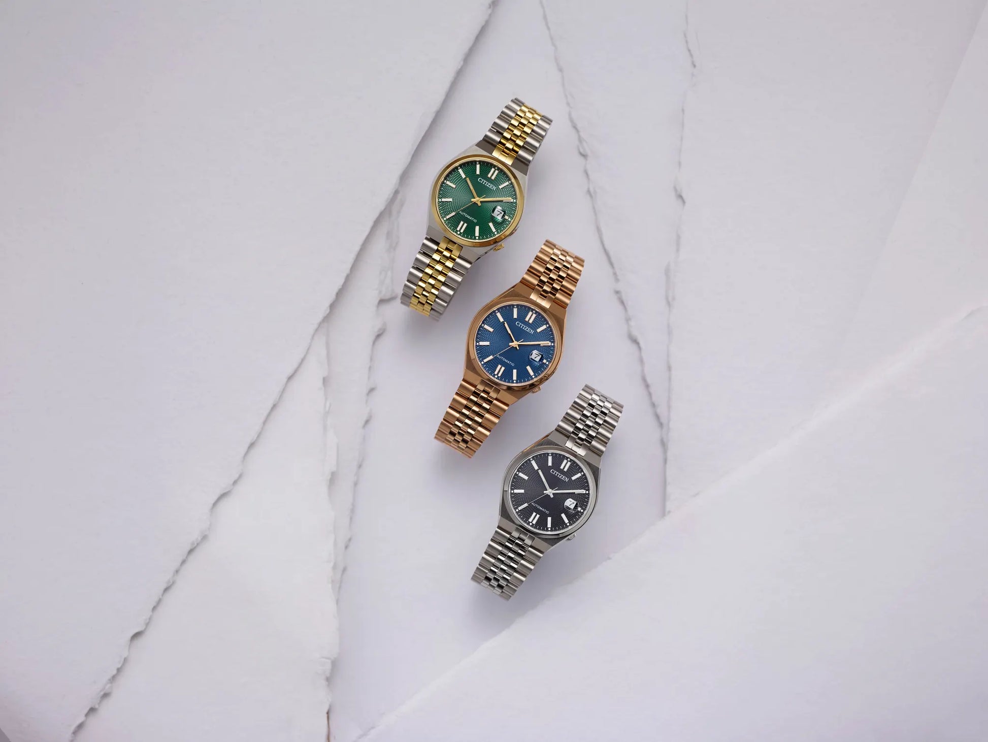 Three women's watch bracelets arranged diagonally on a white marble surface. The top watch has a gold and silver link bracelet with a green dial.