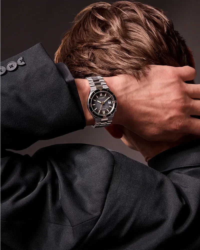 A silver stainless steel wristwatch with a black dial and bezel, worn on a man's wrist. The man is shown from behind, wearing a black suit jacket, with his hand resting on the back of his head against a dark gray background.