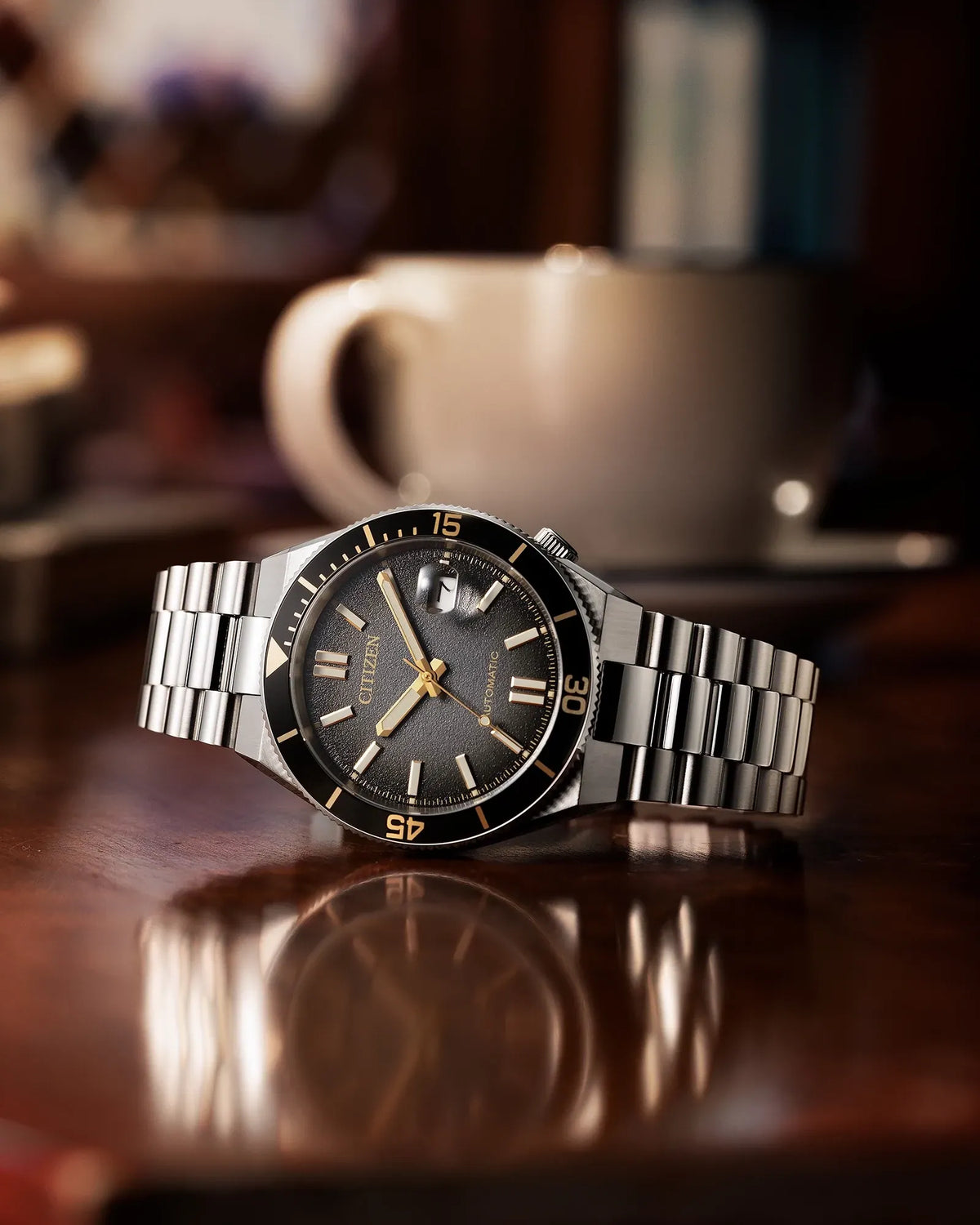 A stainless steel wristwatch with a dark gray dial and black bezel, resting on a polished wooden table with a blurred coffee cup in the background.