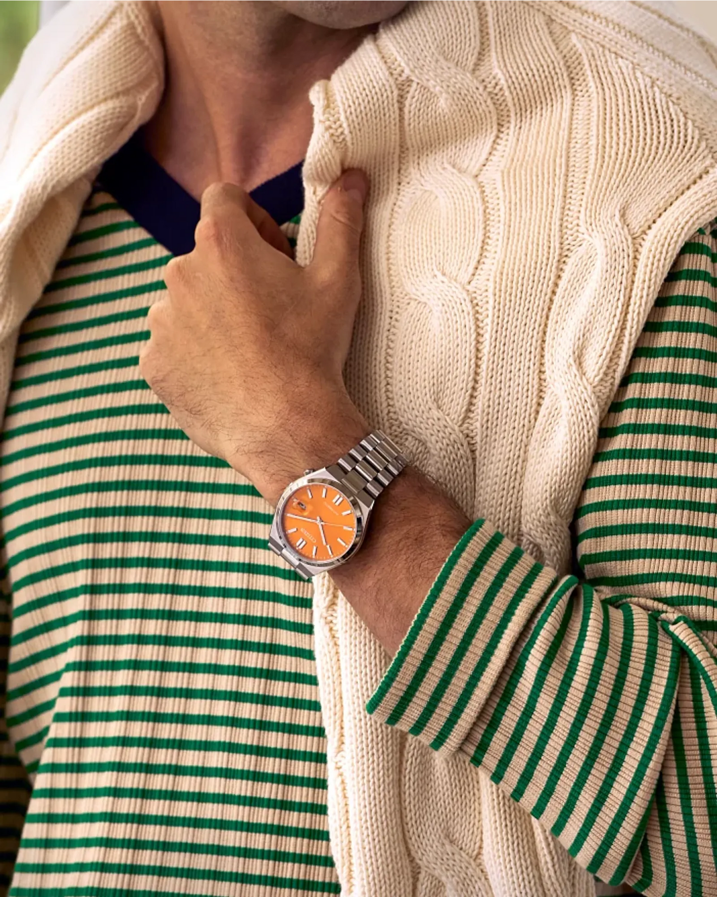 Person wearing a green and white striped shirt with a beige cable knit sweater, adjusting a watch on their wrist.