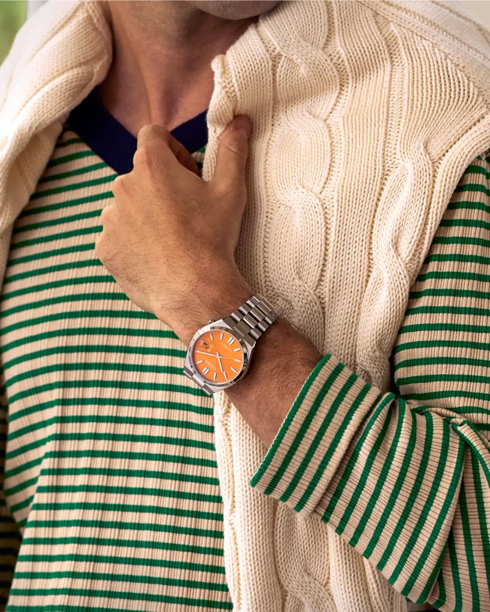 Person wearing a green and white striped shirt with a beige cable knit sweater, adjusting a watch on their wrist.