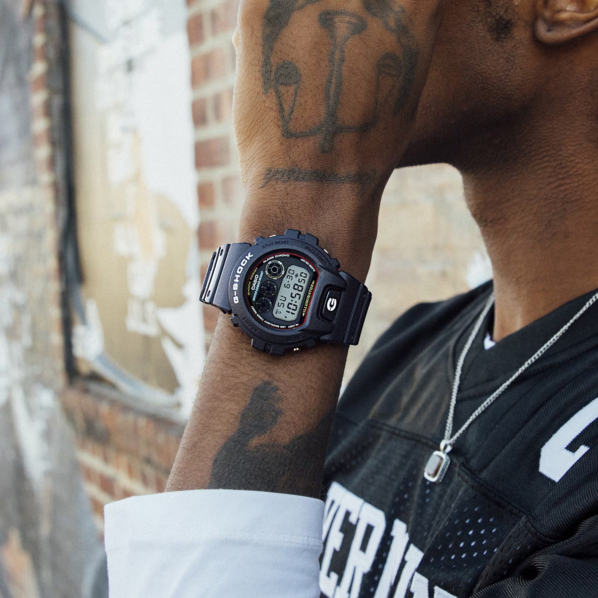 Person wearing a Casio G-Shock watch with tattoos on their arm, standing against a blurred urban background.