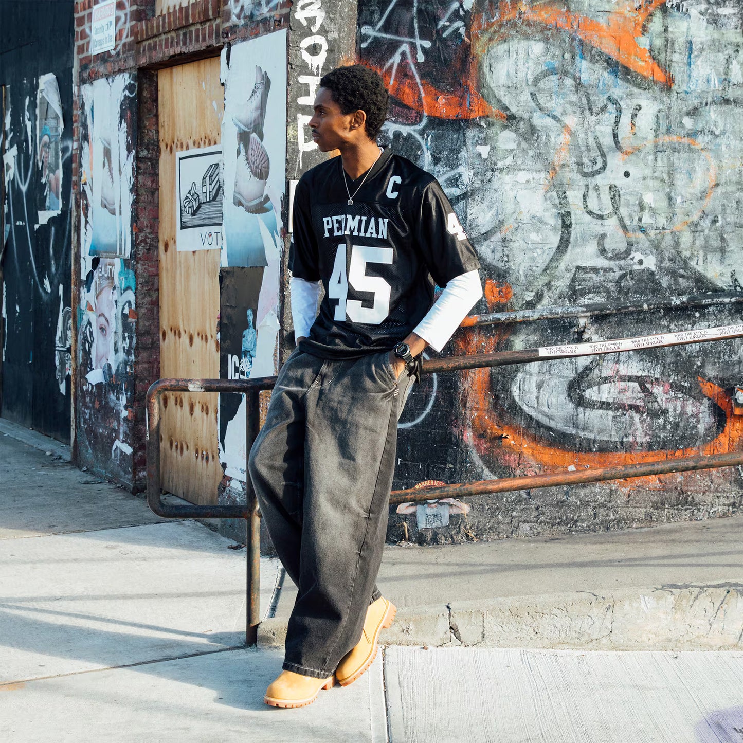 Person wearing a black jersey with white text and numbers, standing against a graffiti-covered wall.