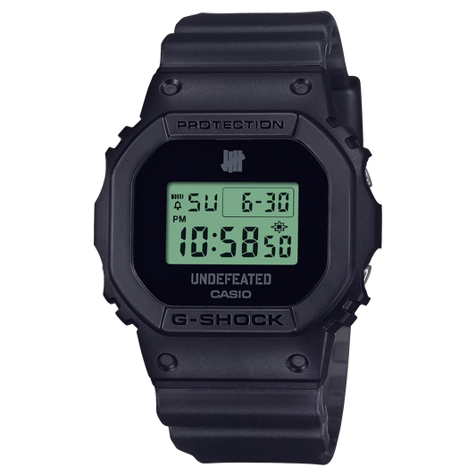 Casio G-SHOCK DWE5600UD-1 UNDEFEATED collaboration model with matte black Carbon Core Guard case, semi-translucent resin band featuring UNDEFEATED logo, green LCD display, engraved caseback, and special packaging. Includes two interchangeable camouflage straps (tiger camo, storm camo). Features shock resistance, mineral glass, 200m water resistance, LED Super Illuminator backlight, stopwatch, countdown timer, alarms, flash alert, auto-calendar, and ±15 sec/month accuracy. Case size 48.9 × 43.8 × 13.7 mm, we