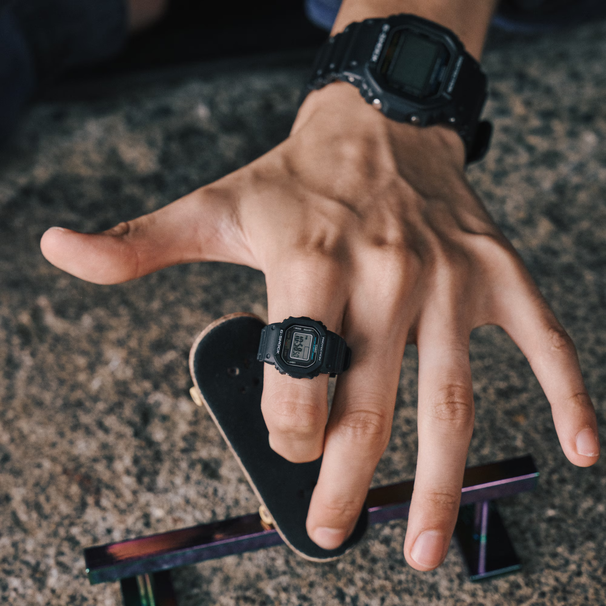 Hand wearing a black digital watch with a small skateboard on a textured surface
