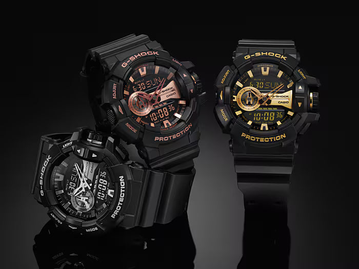 Four G-Shock watches in black and gold on a reflective black surface with a dark background.