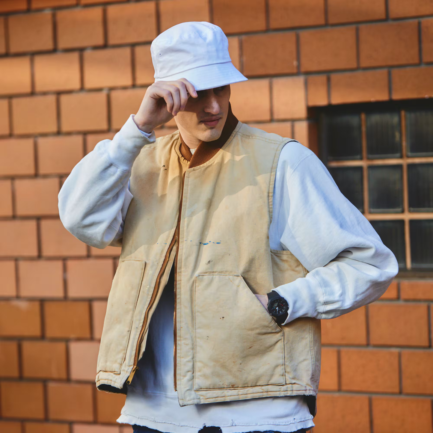 Person wearing a beige vest and white bucket hat against a brick wall.