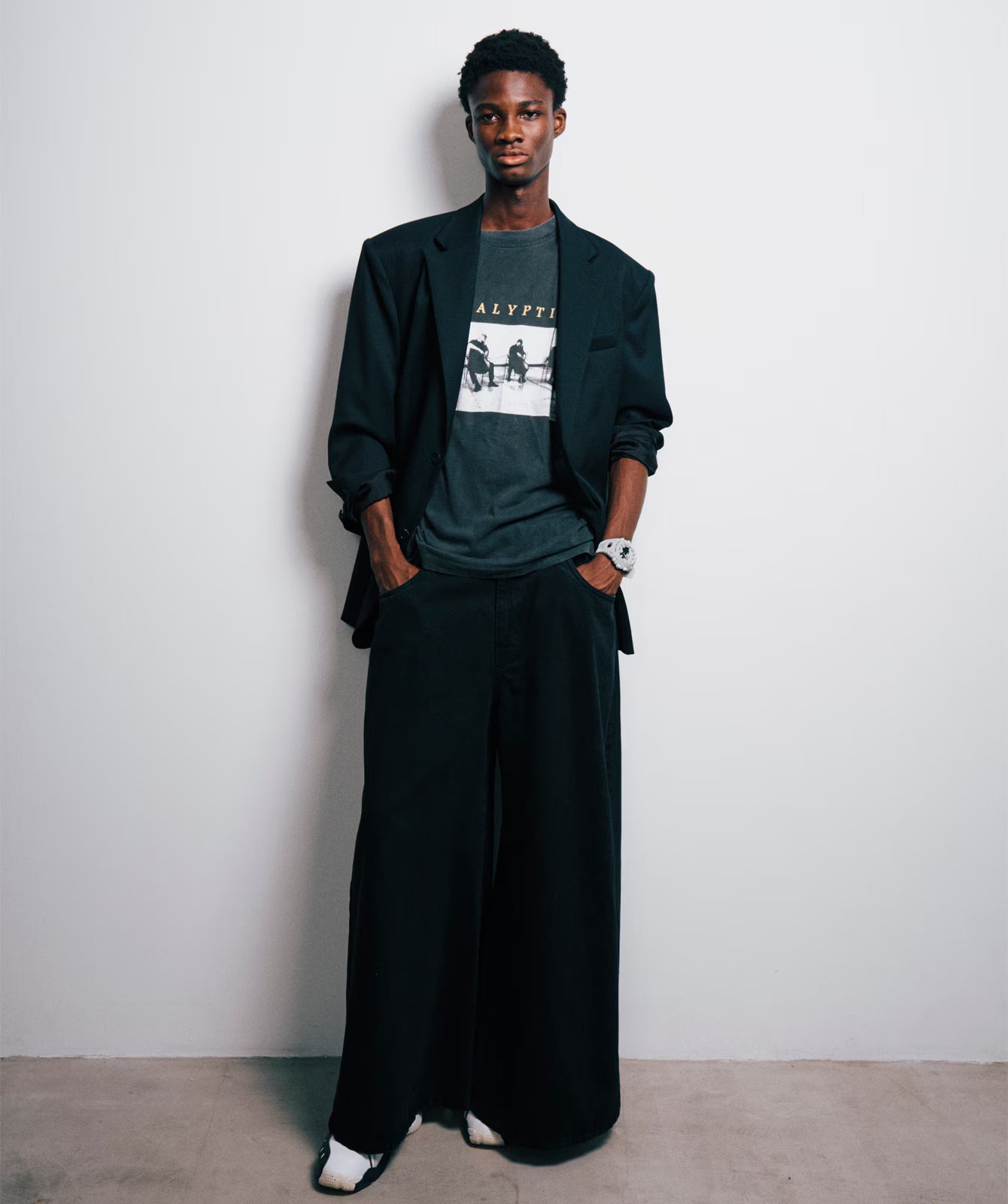 Person wearing a dark blazer over a graphic t-shirt with wide-leg pants against a plain background