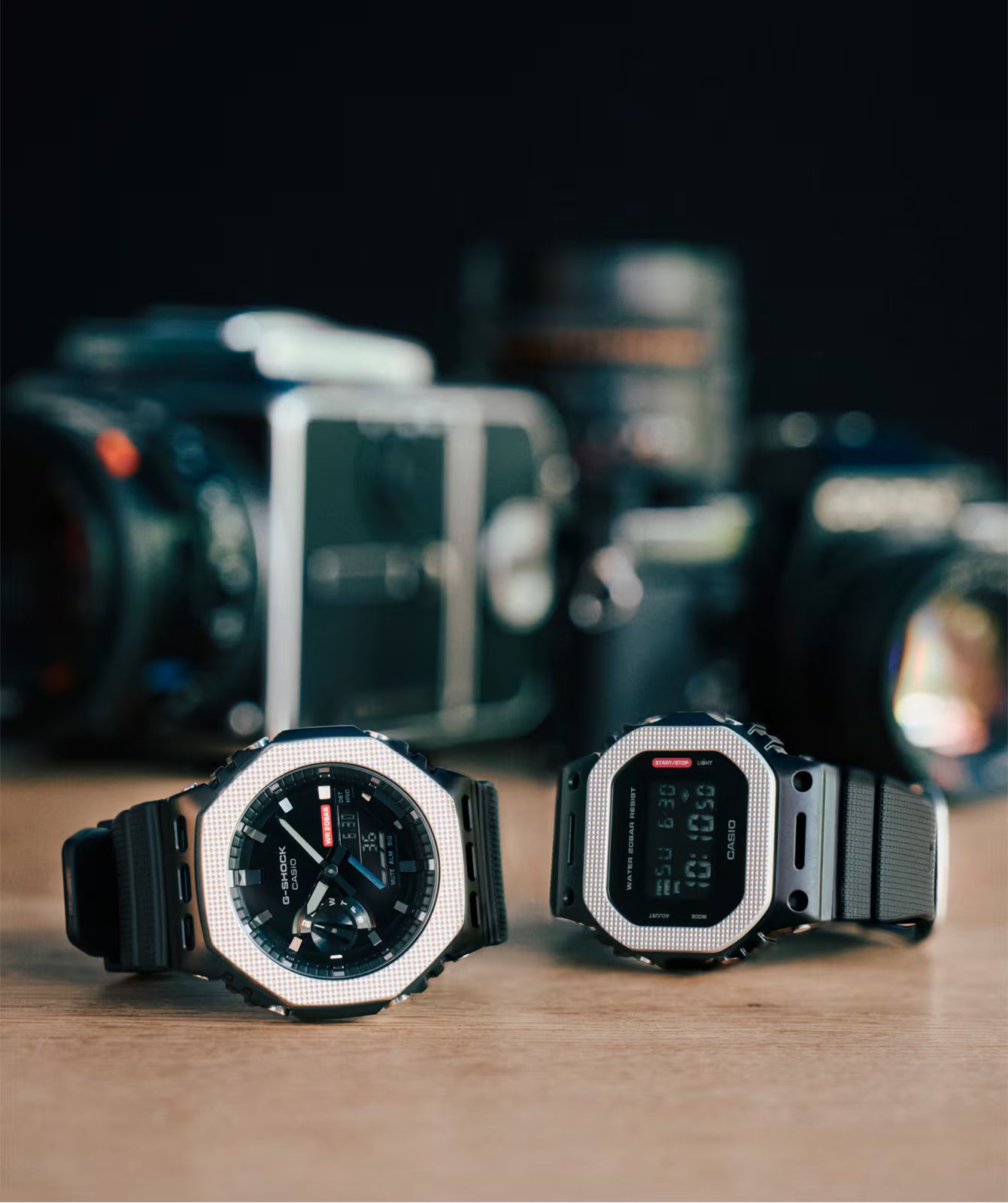Two watches on a wooden surface with blurred camera equipment in the background