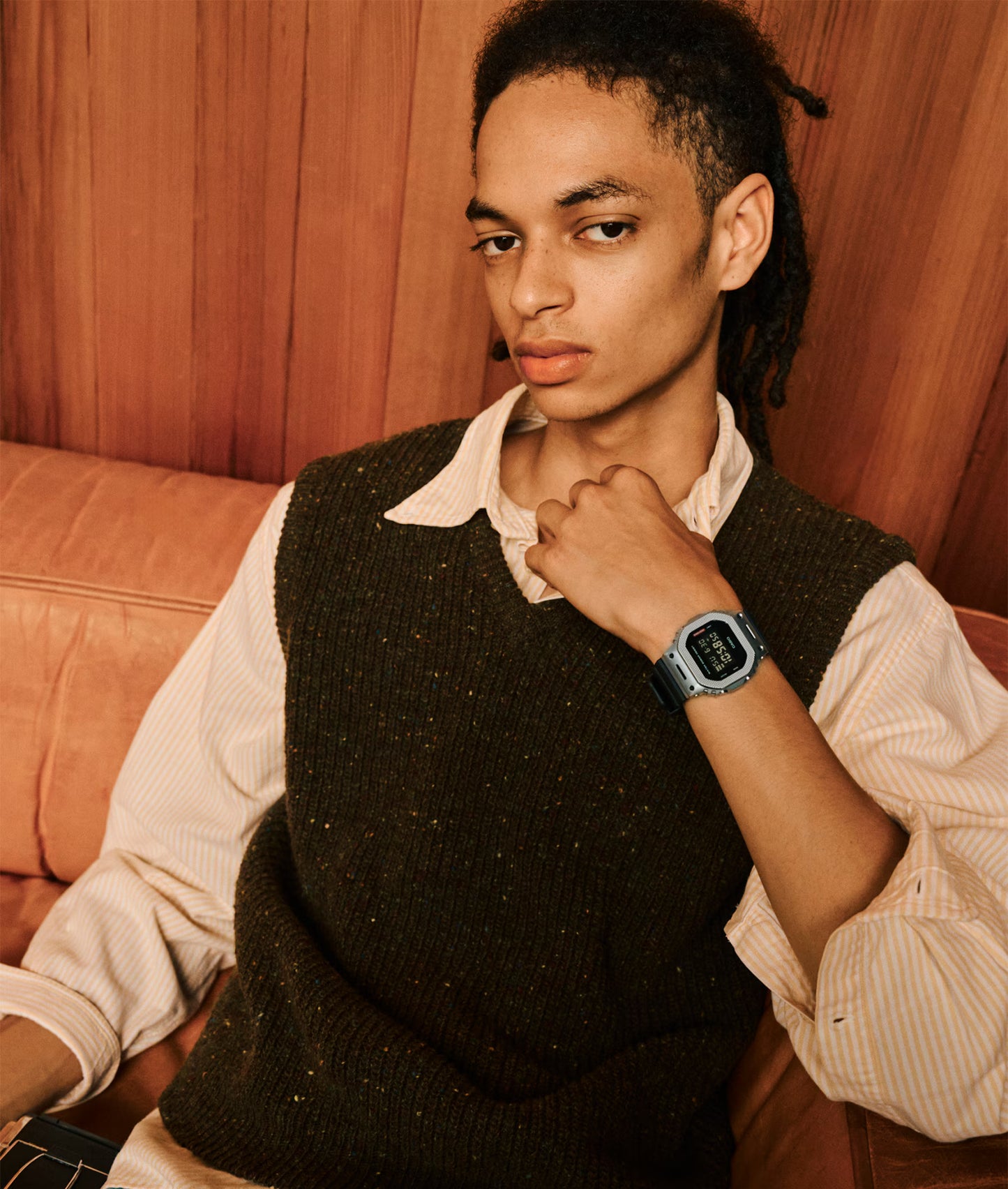 Person wearing a dark sweater vest over a light shirt, sitting on a brown couch with a wooden wall background.