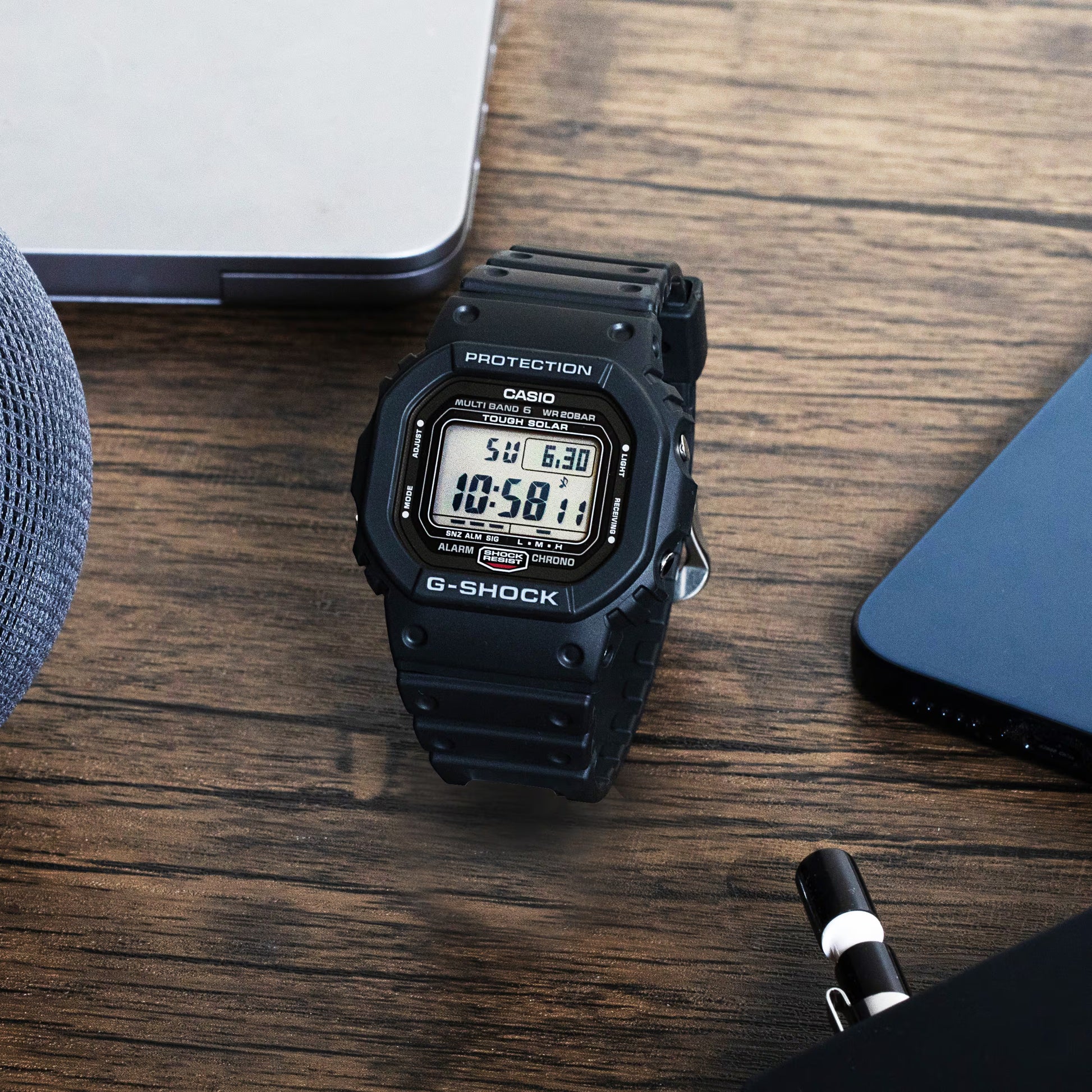 Black Casio G-Shock digital watch on a wooden surface with a pen and notebook.