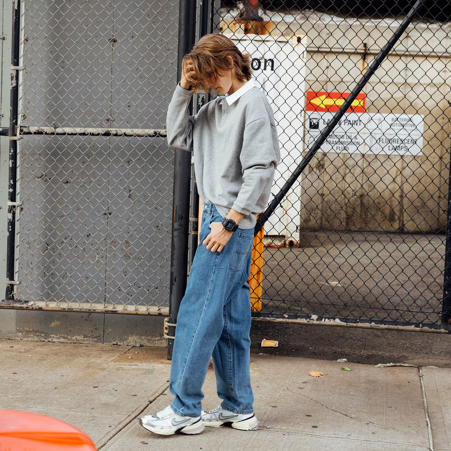 Person wearing a gray sweater and blue jeans standing against a chain-link fence.