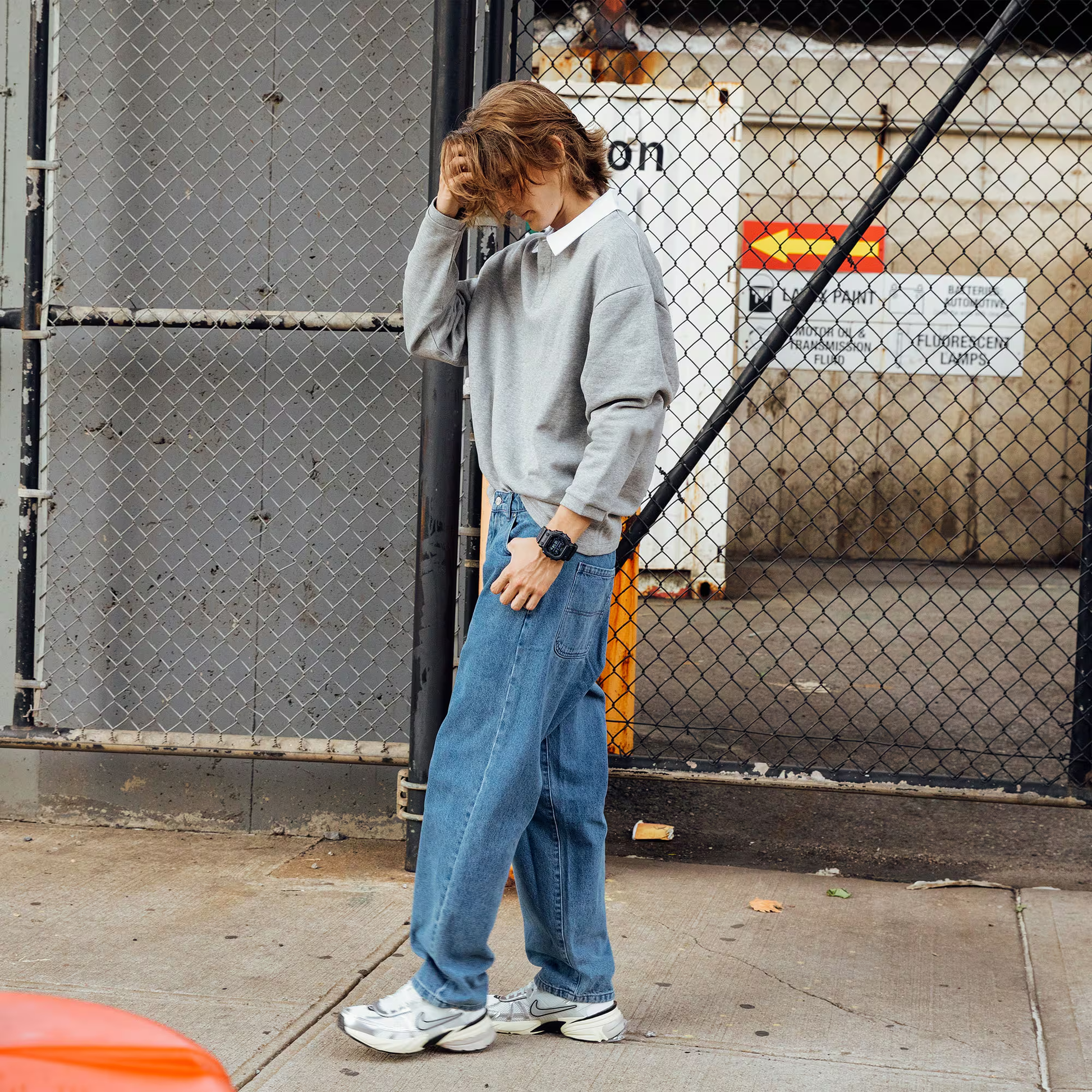 Person wearing a gray sweater and blue jeans standing against a chain-link fence.