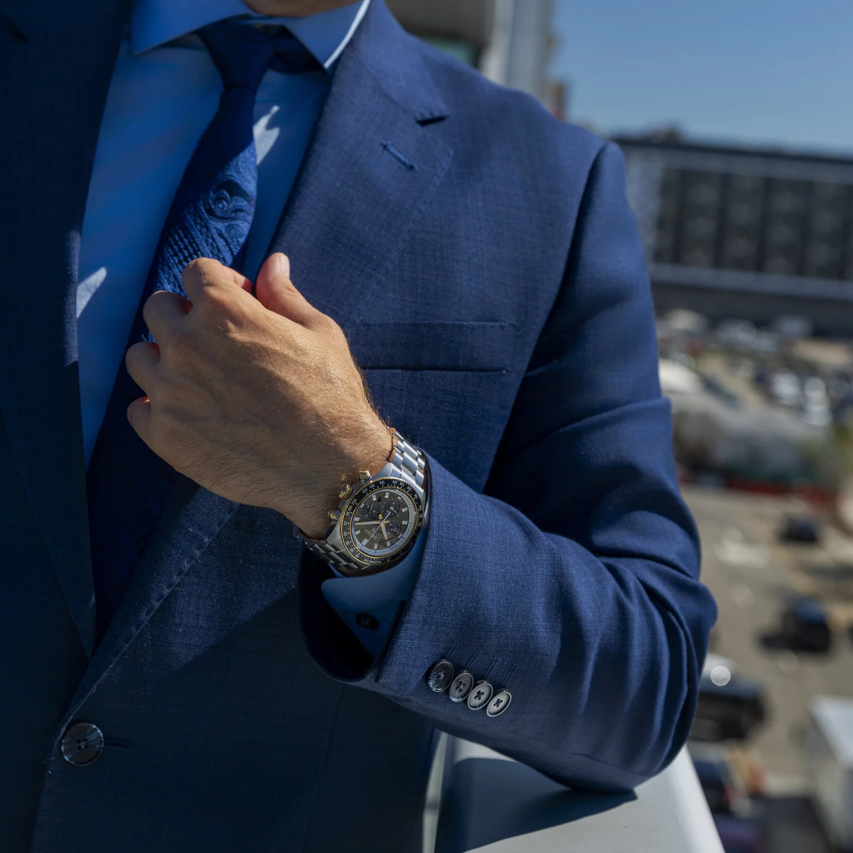 A close-up lifestyle shot of a man in a navy blue suit and tie adjusting his tie while wearing a silver-toned metal watch with a dark dial on his wrist. The background is a softly blurred outdoor urban setting.