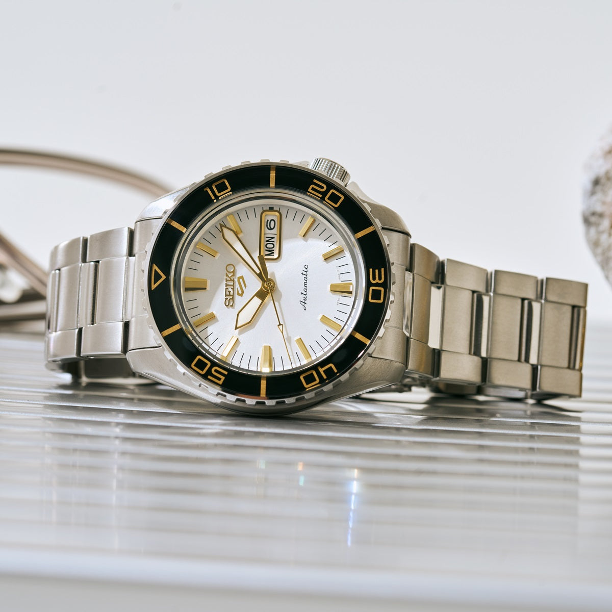 SEIKO 5 Sports HDB004 automatic watch with white dial and black bezel styled flat on a reflective surface with stainless steel bracelet.